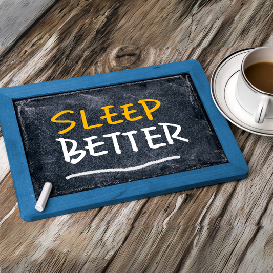 A chalkboard with the text "SLEEP BETTER" written in yellow and white chalk, placed on a rustic wooden surface next to a cup of coffee or tea