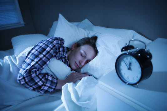 A woman sleeps peacefully cuddled under white sheets on a bed, an alarm clock nearby indicating it's late at night or early morning