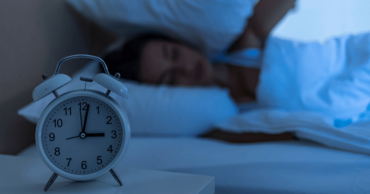A close-up of an alarm clock showing the time while a woman in bed covers her ears, struggling to sleep