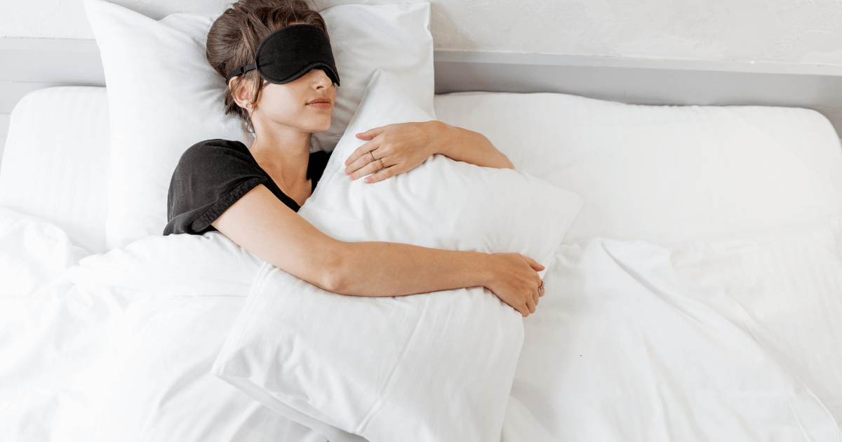 Woman sleeping on her back in bed while hugging a pillow and wearing a black sleep mask | Sip2Sleep®