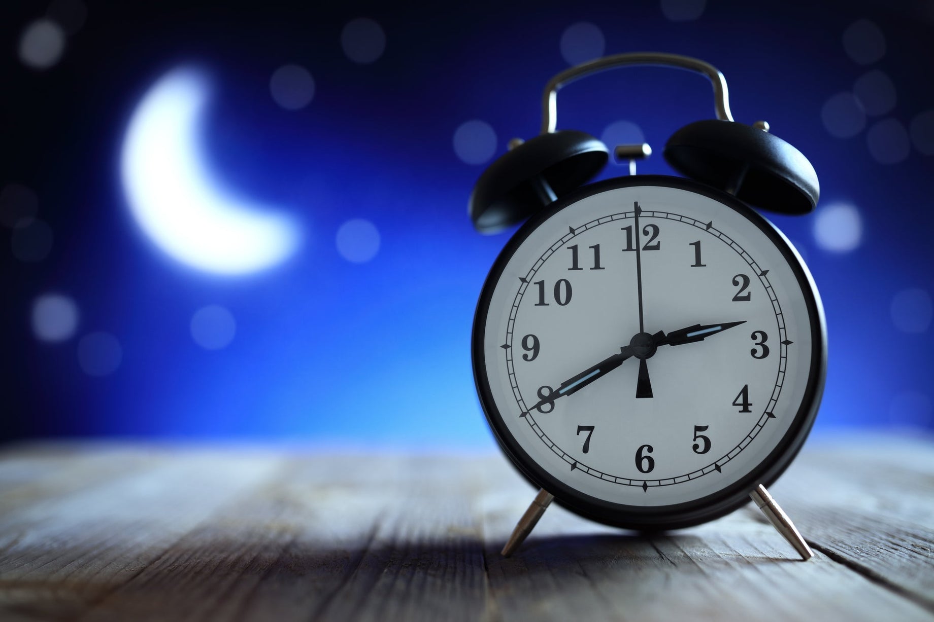 Black alarm clock showing 3 AM on a wooden surface with a blurred night sky and crescent moon in the background, symbolizing broken sleep and nighttime awakenings