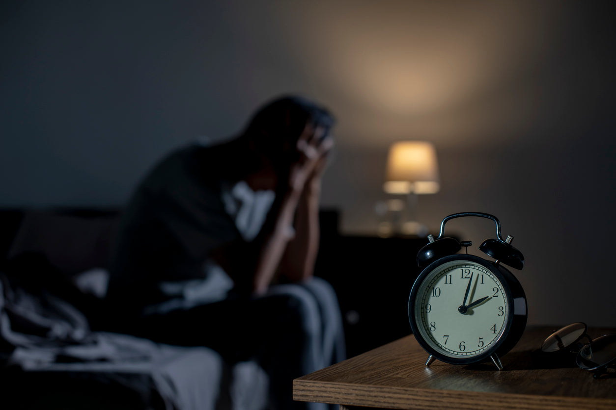 A dimly-lit bedroom scene shows a person sitting on the edge of a bed, head in hands, appearing distressed or fatigued despite the late hour shown on the alarm clock nearby. 