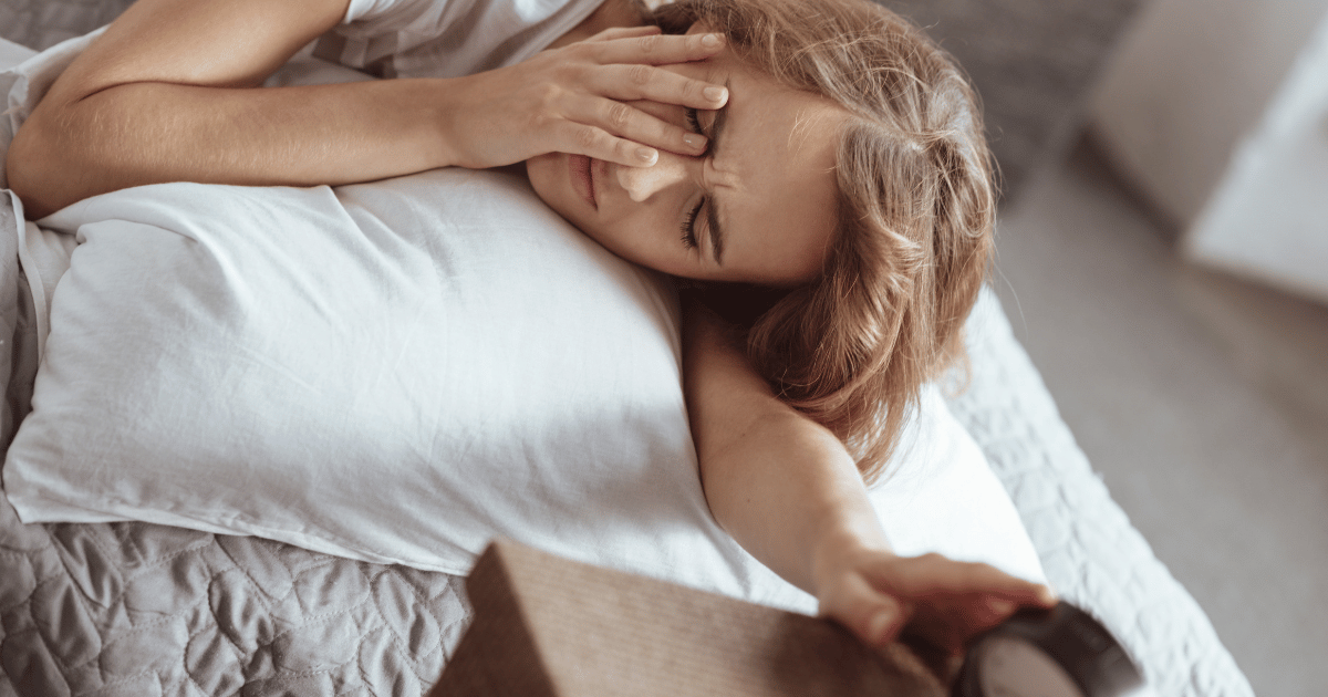 Woman waking up groggy, covering her eyes and reaching for her alarm clock