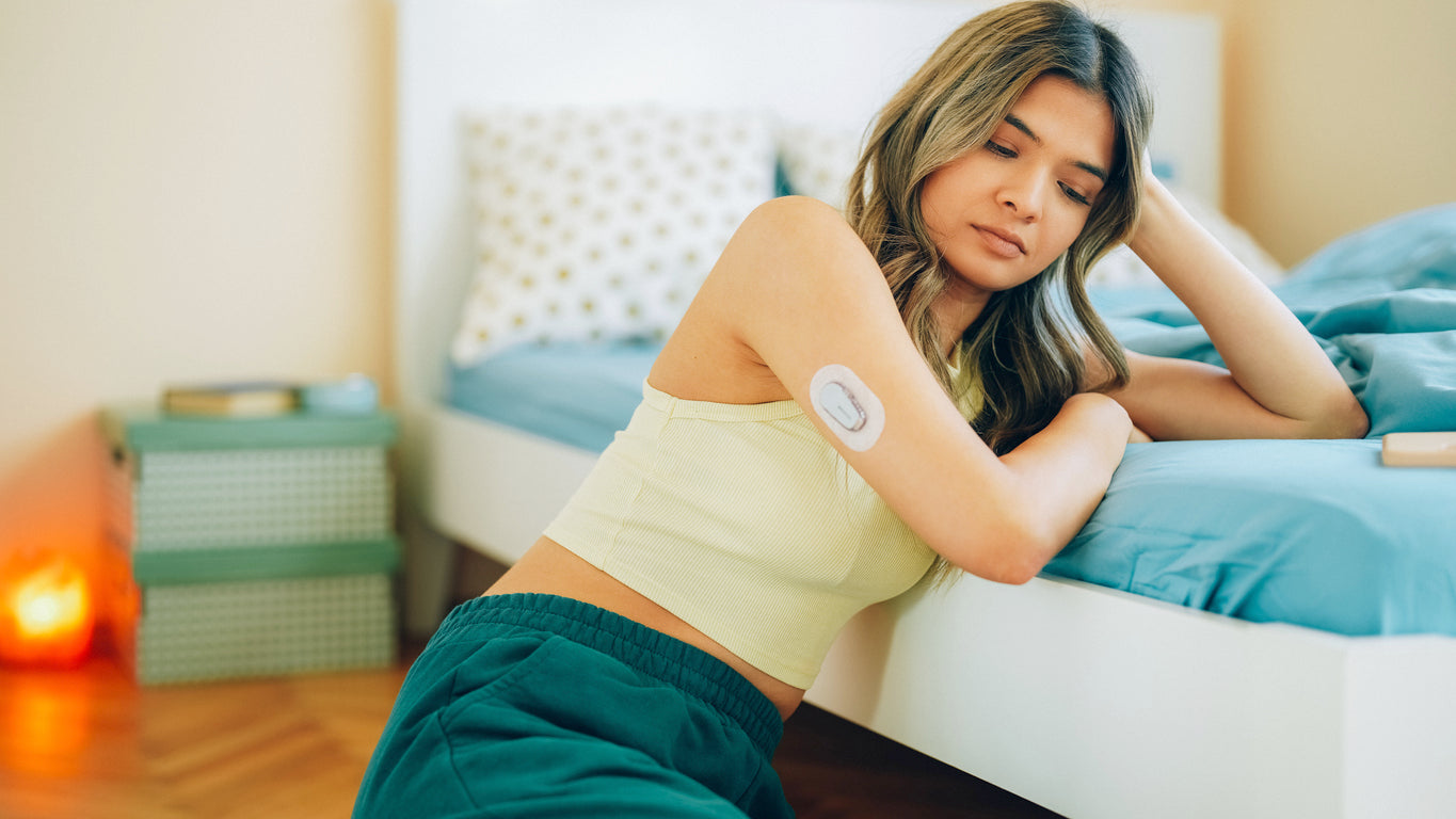 A young woman with long brown hair sits thoughtfully on her bed, resting her head in her hand, appearing pensive about managing her blood sugar levels through proper sleep habits