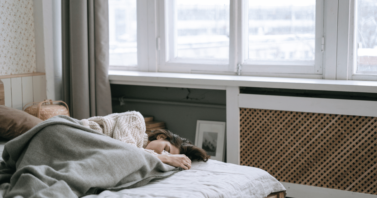 A woman sleeping in bed near a window with natural light coming through, illustrating how seasonal daylight changes can affect sleep