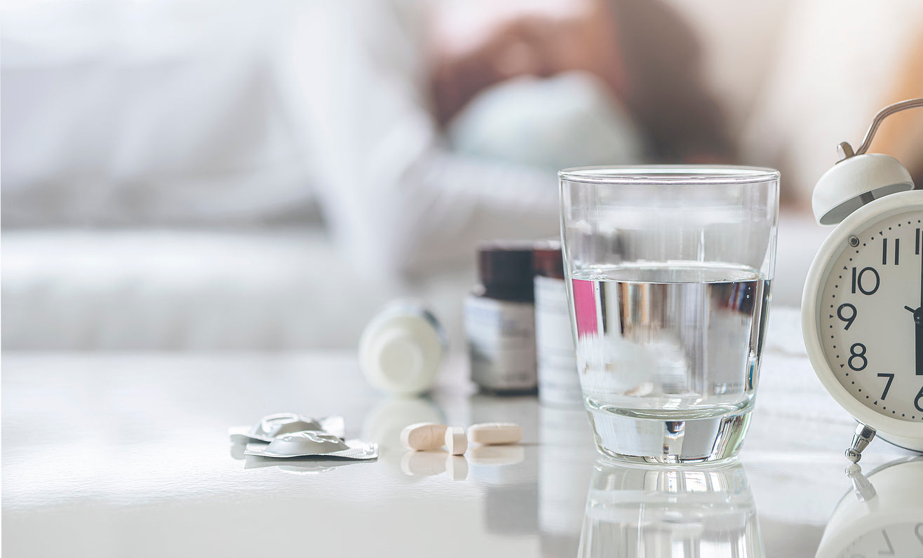 Bedside table with a glass of water, pills, medicine bottles, and an alarm clock. A person is visible in the background, sleeping in bed. The image suggests various non-prescription sleep aids and remedies