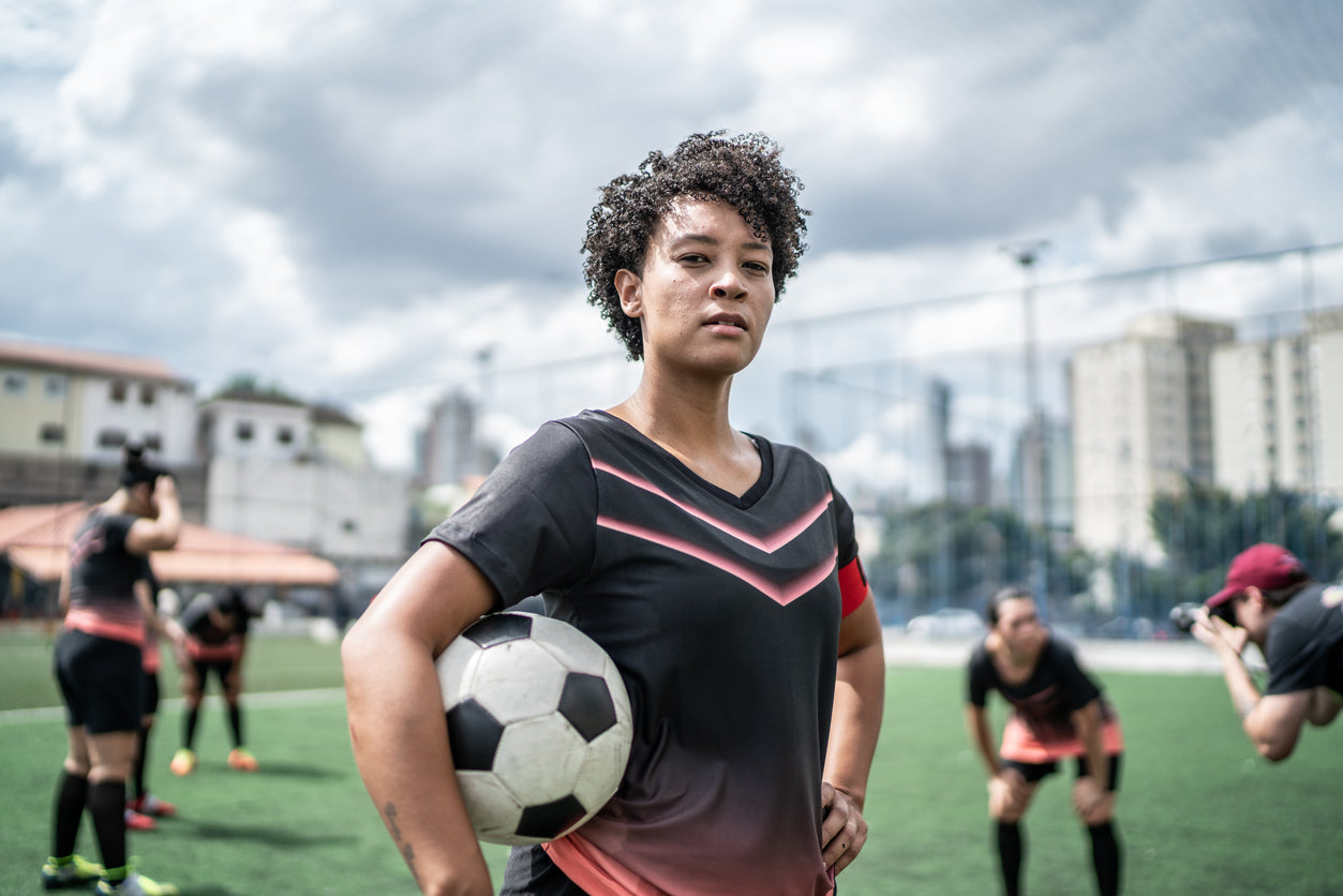 a young athlete with curly hair holding a soccer ball, standing on a field with other players practicing in the background. The athlete has a determined expression, suggesting the importance of proper rest and recovery for optimal athletic performance