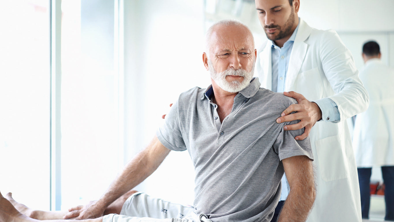 Senior man grimacing in pain while seated, with a doctor's hand on his shoulder offering support. The scene suggests discomfort possibly related to inflammation, highlighting the connection between physical health and sleep quality